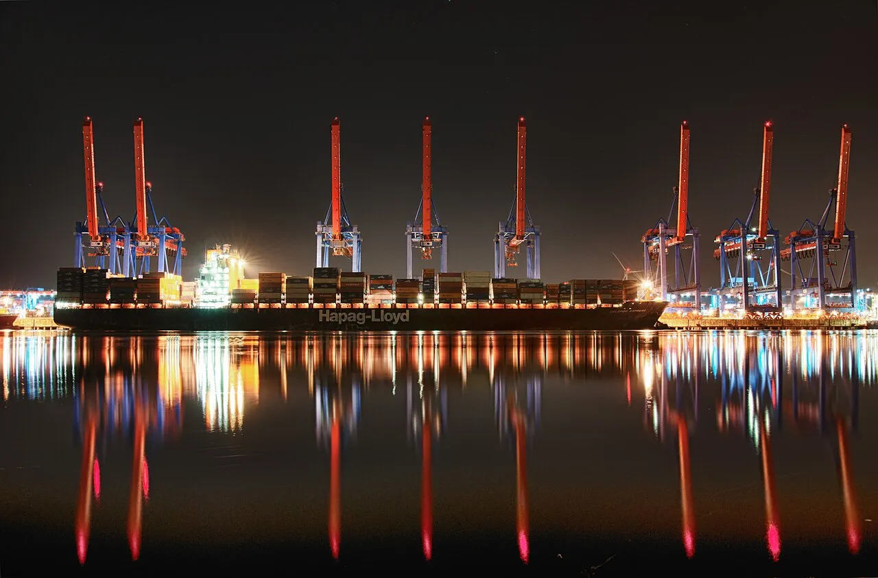 Gantry cranes lit red over a container ship at night, reflected in the river